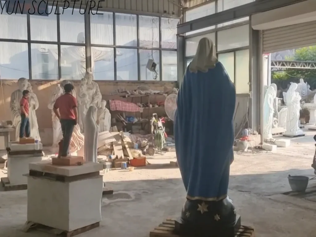 The back of the statue of the Virgin Mary, photographed in the Cloud Sculpture workshop. The deep blue shawl showcases an exceptionally natural sculpting technique that captures the fabric’s drape, while the star-shaped decorative motifs on the base reveal meticulous casting details. In the background, the statue of St. Bernadette is shown undergoing a scale check, illustrating the preparatory phase for the visual alignment of the two figures within the grotto setting.