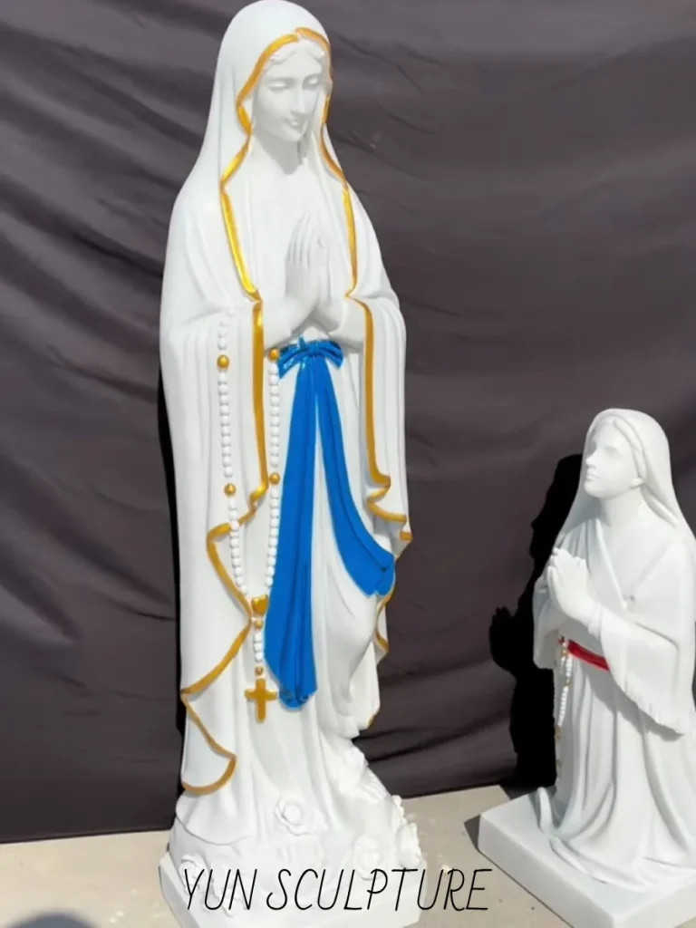 A masterful white marble our lady of lourdes and bernadette statue composition standing in a workshop. The young saint kneels below in historically authentic peasant attire, her hands clasped in prayer with her gaze fixed sharply upward. This precise relational narrative perfectly captures the critical "Line of Sight" calculation, ensuring the invisible intersecting gaze between the divine apparition and the mortal witness preserves the sacred intimacy of the 1858 encounter.