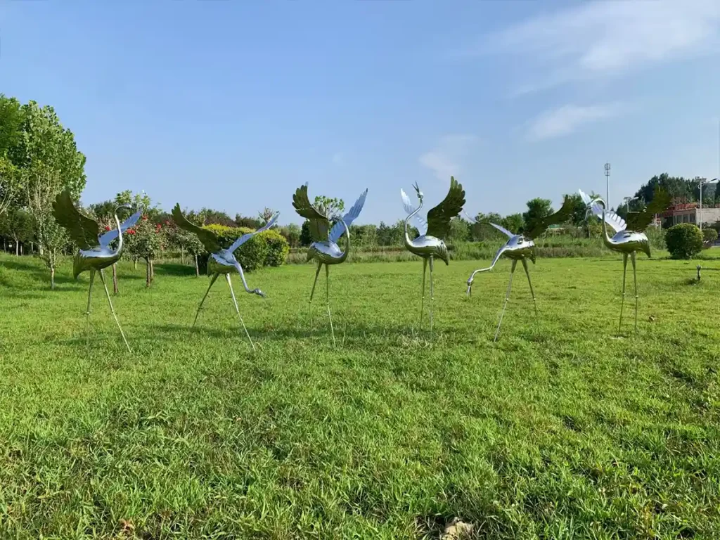 A stunning group of outdoor metal animal sculptures featuring elegant cranes on a lush green lawn. Each mirror polished stainless steel sculpture flawlessly reflects the surrounding landscape and blue sky, demonstrating the extraordinary visual dynamism of contemporary metal art.