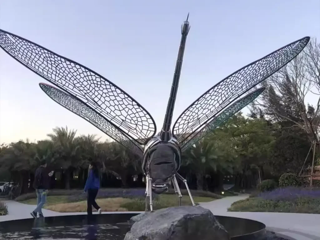 An impressive outdoor metal dragonfly sculpture appearing to hover over a shallow reflective pool in a public park. The highly reflective stainless steel body captures the surrounding light and sky, creating a surprisingly strong visual impact for a fantasy-themed landscape.