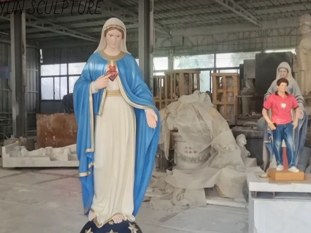 This close-up of the Virgin Mary statue showcases Cloud Sculpture’s signature hand-painted craftsmanship. The icon’s facial expression blends compassion with a sense of youthfulness, reflecting St. Bernadette’s description of the “Beautiful Lady.” The blue-and-white robe, accented with gold trim, demonstrates exceptional artistic quality. A scale model in modern attire is visible on the right, used to ensure the visual balance of the large-scale icon within the public landscape. Translated with DeepL.com (free version)