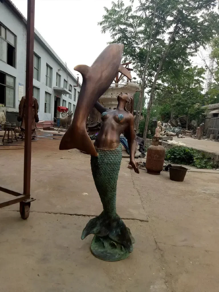 A beautiful large bronze mermaid sculpture undergoing the patination process in a workshop yard. The striking dual-tone finish—a warm brown human torso and a sea-green textured tail—perfectly symbolizes the meeting of two worlds, land and sea, embodying the mystery of the ocean spirit.