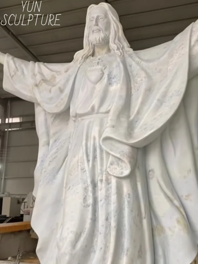 A frontal close-up of a beautiful white stone statue of Jesus revealing subtle natural grey veining across the torso. These beautiful but porous natural lines act as micro-fractures; if placed outdoors, water infiltrates and freezes within them, proving that while beautiful, stone is too brittle for extreme outdoor church landscapes.
