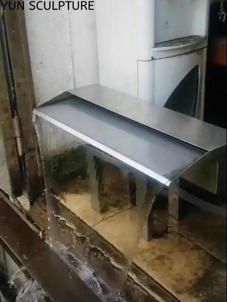 Extreme close-up of a precision-machined stainless steel waterfall weir producing a flawless, glass-like sheet of falling water during a factory flow test. This critical engineering detail ensures the continuous rain curtain effect, allowing light to graze the fluid surface and amplify the depth of the modern landscape design.