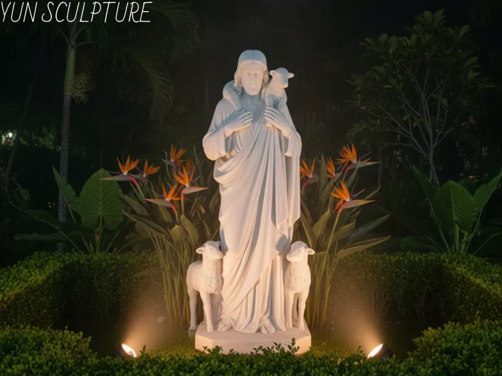 An illuminated white marble statue of Jesus as the Good Shepherd, holding a lamb and standing in a private estate healing garden at night. The sculpture is lit by two spotlights, creating a peaceful, intimate ambiance. Bird of Paradise flowers and dense tropical foliage surround the base, showcasing high-quality custom stone work for spiritual landscapes.