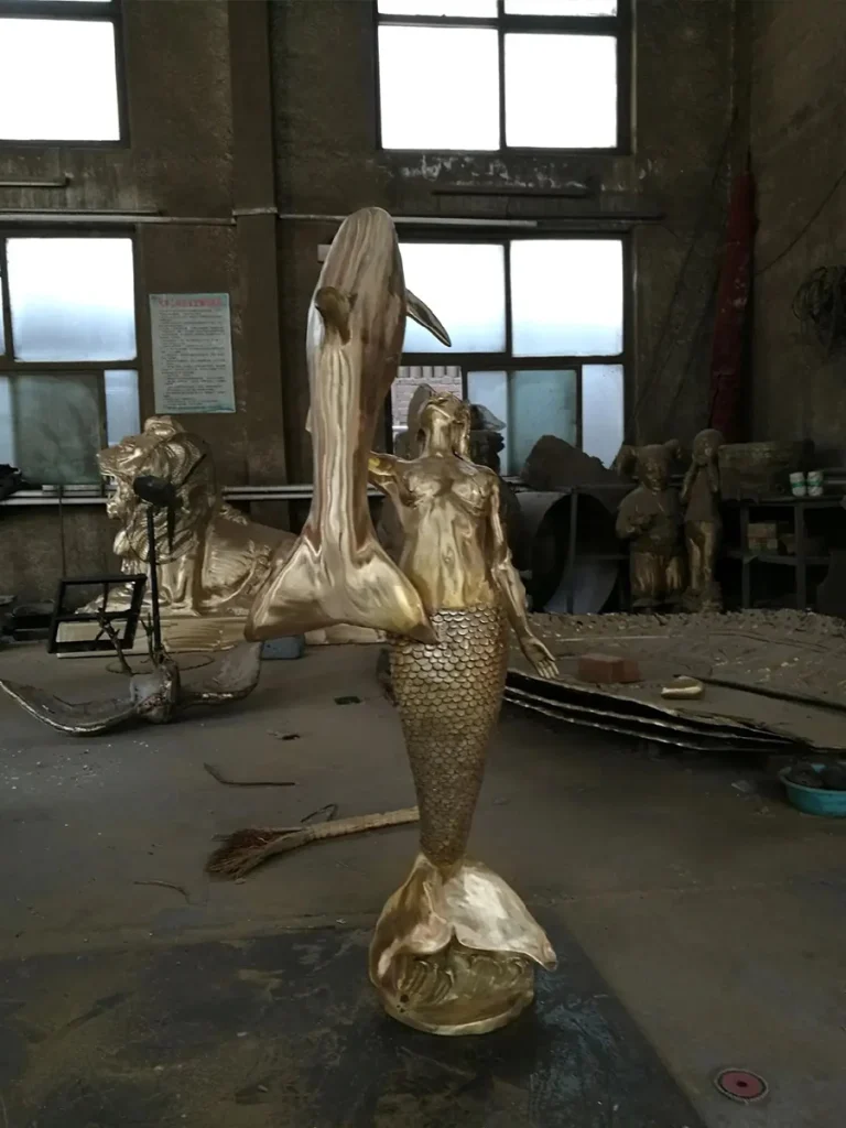 A large, raw-cast bronze mermaid statue holding a leaping dolphin, standing inside a foundry. The gleaming golden metal highlights the sculpture's elegant vertical movement, capturing the dynamic energy required to become the visual centerpiece of a luxury poolscape.