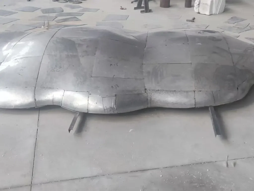 The initial fabrication stage of a large luxury stainless steel sculpture. Assembled from numerous cut steel plates, the structure displays highly visible weld seams, illustrating the raw starting point before master craftsmen begin the meticulous finishing process.