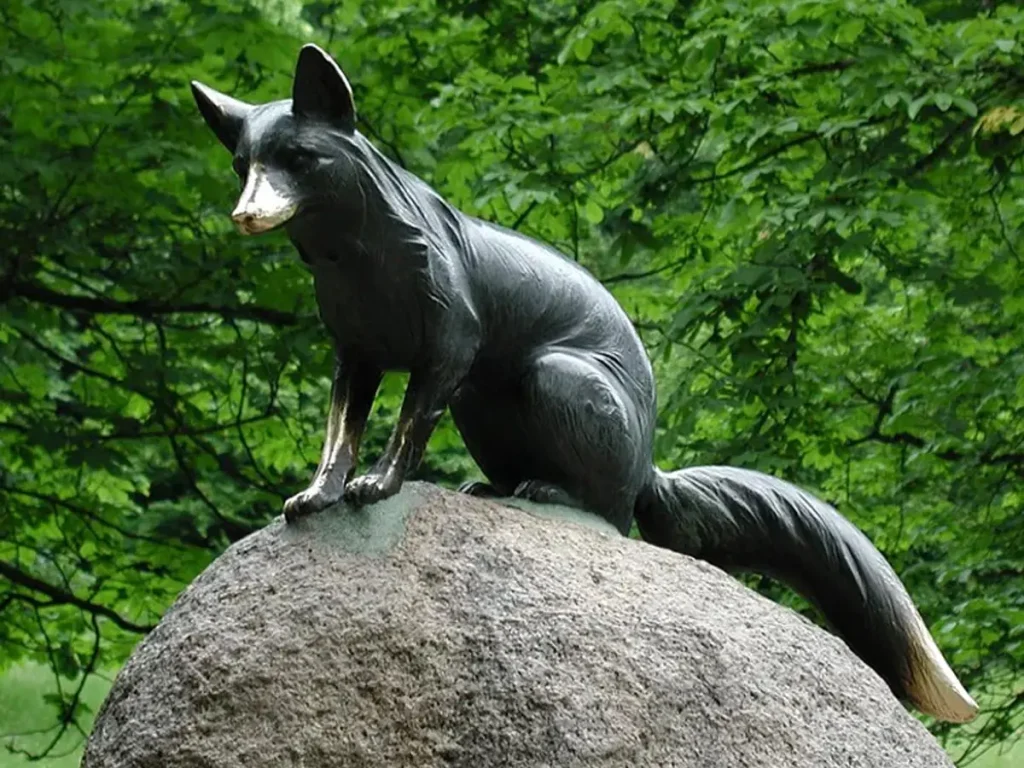 A highly detailed dark bronze fox sculpture perched gracefully atop a large natural boulder against lush green foliage. This strategic placement blends the fine metal art with the natural textures of stone and leaves, bringing the forest-themed garden landscape to life.