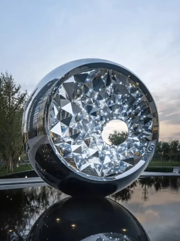 A mesmerizing geometric sculpture crafted from polished stainless steel, transforming a modern plaza into a memorable landmark. The crystal-like hollow center and the smooth outer shell brilliantly capture the movement of light and sky reflections, inviting interpretation without dictating a specific meaning.