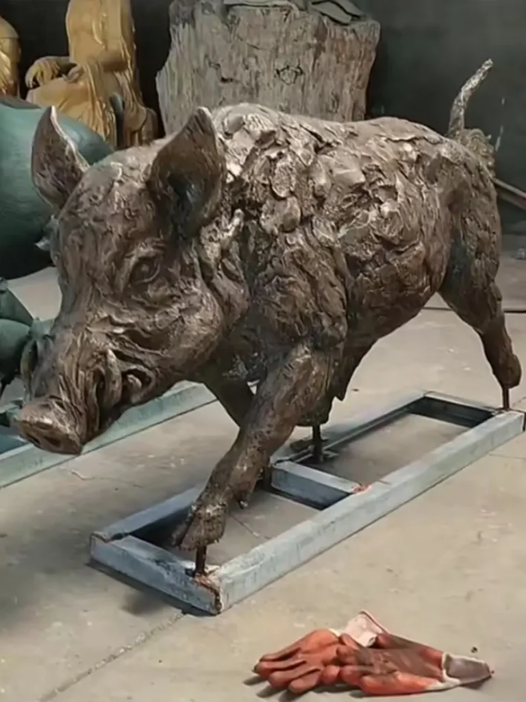 A highly textured bronze wild boar statue mounted on a metal frame in a workshop. The rugged, realistic fur and fierce forward motion echo deep European hunting traditions, making it an ideal, powerful focal point for a rustic lodge entrance.