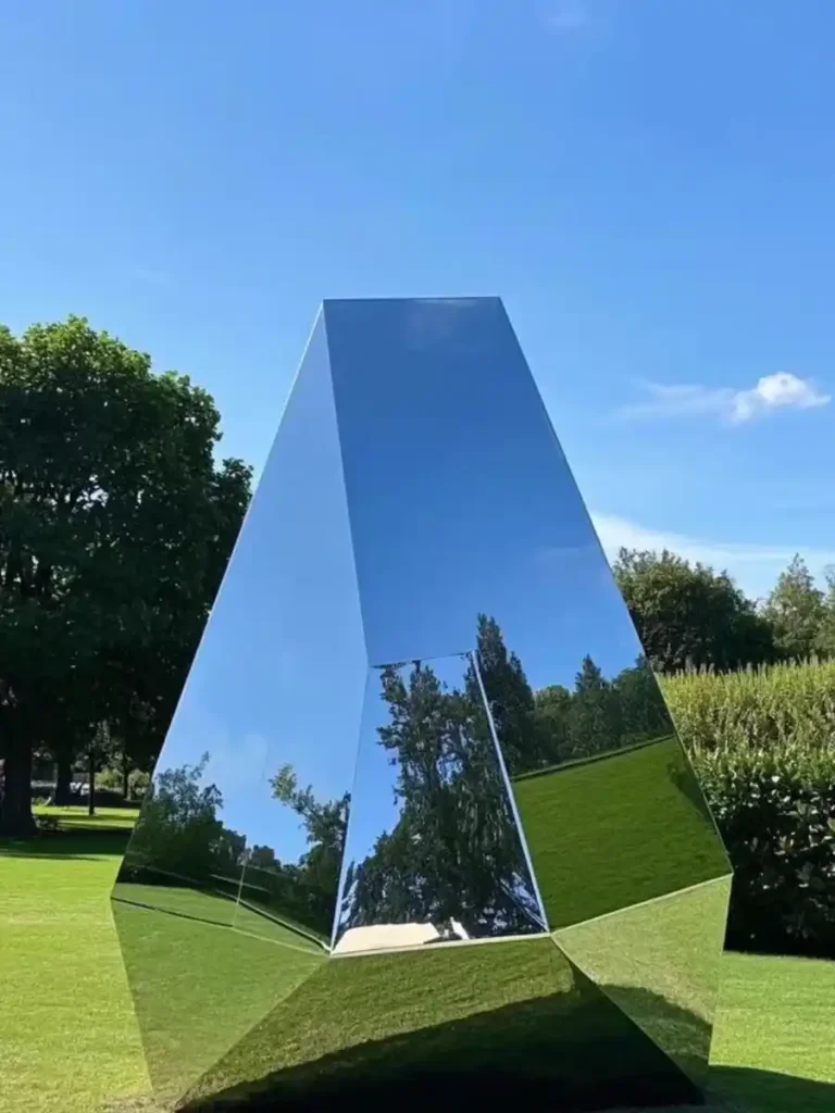 A massive, mirror-polished geometric metal sculpture standing confidently on a lush green lawn. Showcasing safe installation without visible hardware, this flawless contemporary artwork relies on a concealed mounting system and internal anchor plates, ensuring the heavy engineering disappears completely into the landscape.