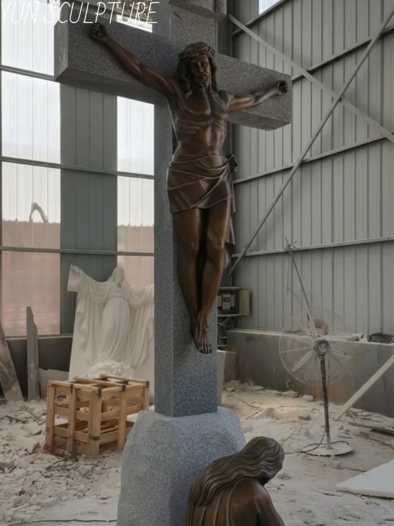 An angled view of a massive bronze crucified Jesus statue attached to a sturdy stone-finish cross, with Mary kneeling at the base. This impressive workshop scene highlights how large corpus pieces are expertly cast in sections, ensuring safe transport before being TIG welded and seamlessly patinated on-site onto a heavy-duty internal armature.