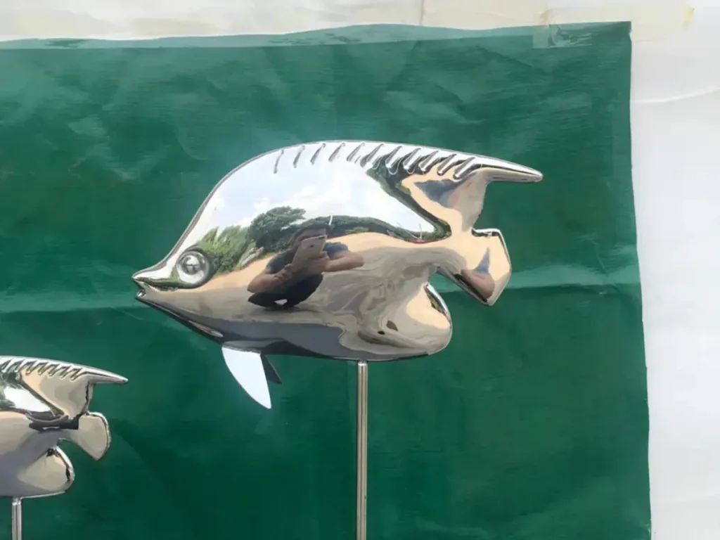 Close-up of a highly polished metal fish sculpture reflecting the surrounding greenery. When placed among stone and water plants, this piece introduces bright, playful motion into an otherwise still garden environment.