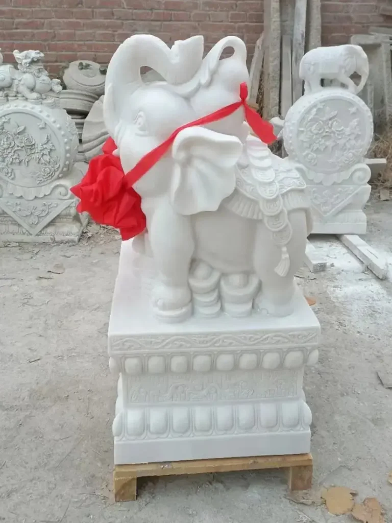 A small, adorable white marble elephant statue with its trunk raised high, decorated with a red bow. Often called a "good luck elephant," this trunk-up design is frequently placed in entryways to invite success, career growth, and prosperity.