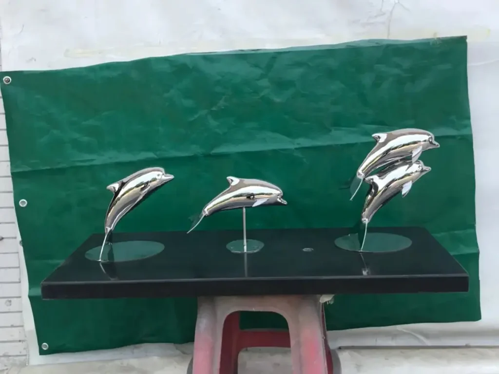 Three miniature mirror-polished stainless steel dolphin sculptures resting on a dark base. These sleek aquatic models capture the playful motion and fluid arcs that make them perfect subjects for luxury resort pool installations.