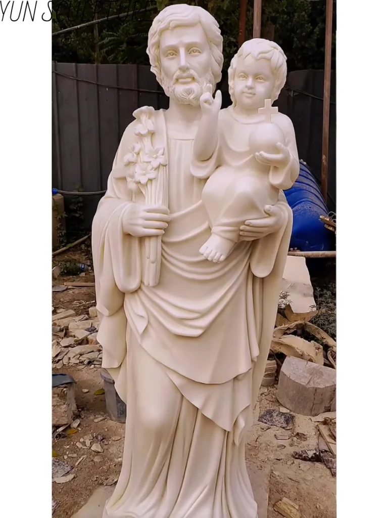 A detailed marble carving of Saint Joseph holding the infant Jesus, showcasing the transition from clay model to finished stone sculpture.