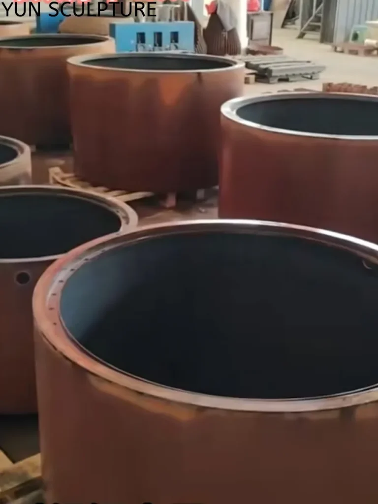 Several nearly finished Corten steel water fountain basins lined up in a warehouse. Featuring a dark, highly stabilized pre-oxidized exterior and a protective black interior coating, this meticulous factory process contains the release of free iron oxide particles, delivering immediate visual maturity and alignment with the architect's design intent from day one.