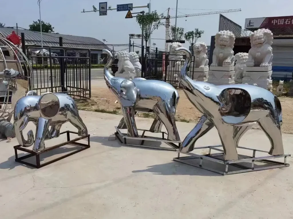 A group of three abstract mirror-polished 316 stainless steel elephant sculptures reflecting the outdoor factory environment. Utilizing modern forging techniques, these smooth, corrosion-resistant statues create a striking and dynamic visual presence for large modern landscape designs.
