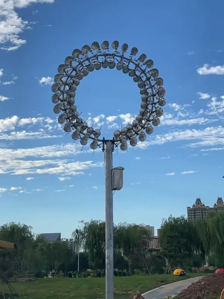A static, compact view of a kinetic stainless steel wind sculpture, showing the first stage where clear reflections of the sky establish the Liquid Silver effect.