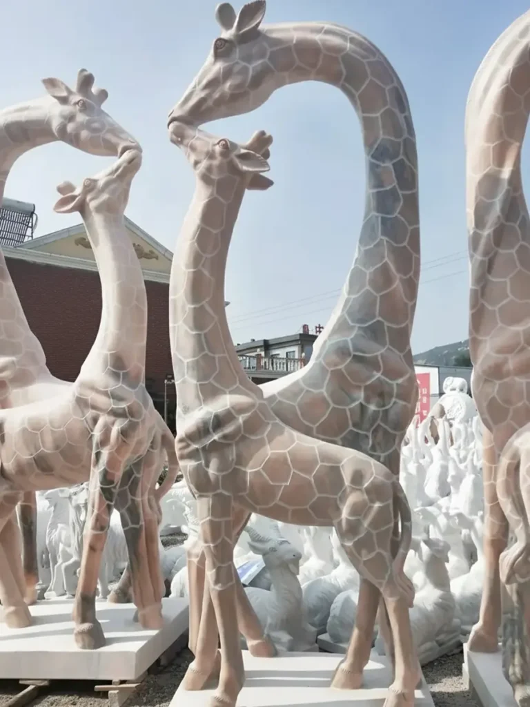 Two beautiful giraffe statues carved from sunset red marble standing in a stone workshop. The taller giraffe gently bends its neck toward the smaller one, perfectly embodying the calm elegance, grace, and elevated awareness described in contemporary garden symbolism.