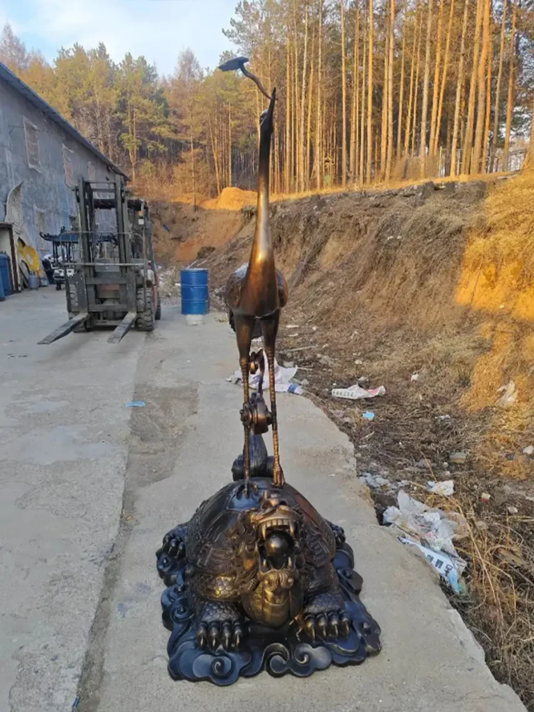 Front view of an elongated bronze crane statue perched on a carved turtle base, bathed in the warm golden hour light of a factory yard. The sculpture's warm patina and strong vertical form effortlessly bring Zen philosophy directly into any landscape design.