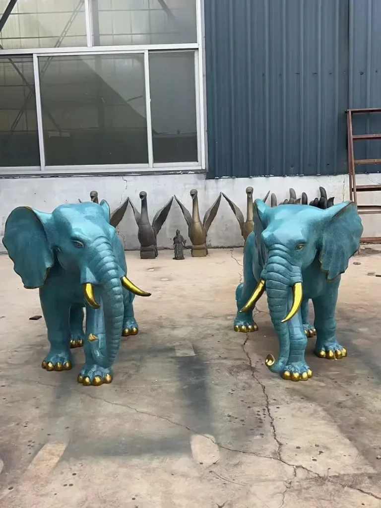 A pair of teal-patina bronze elephant statues with golden tusks standing in a factory, their trunks lowered toward the ground. This downward posture represents the elephant's natural behavior of interacting with the earth, symbolizing fertility, longevity, and grounded strength.