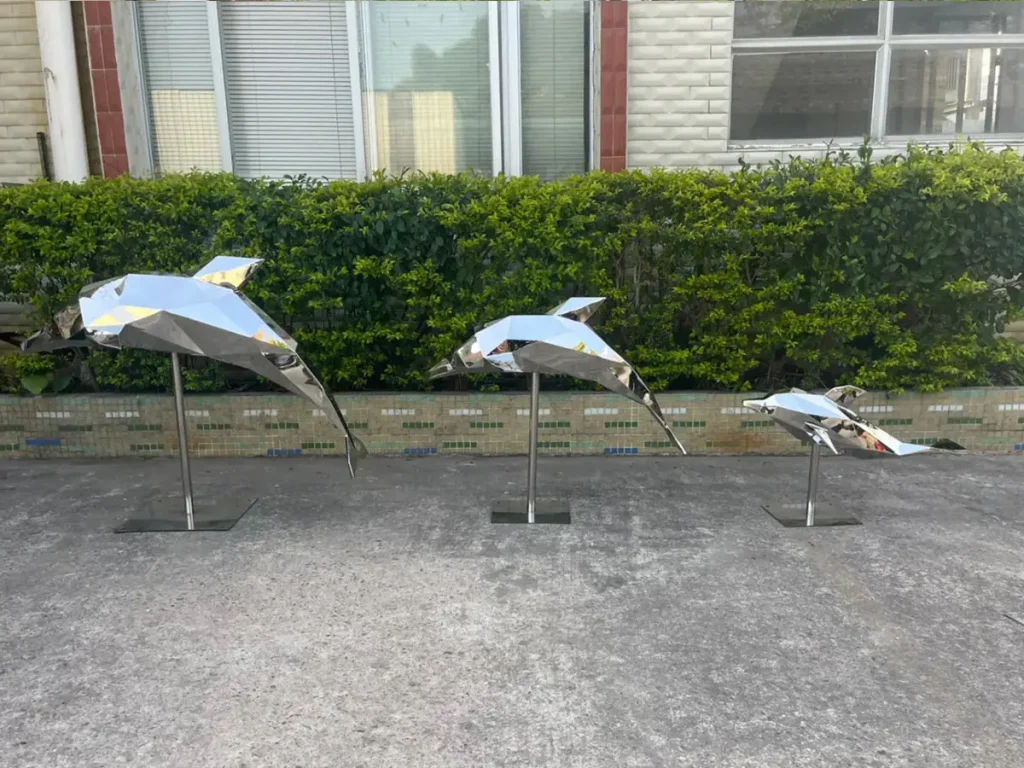 Three geometric mirror stainless steel dolphin sculptures mounted on stands, reflecting their outdoor surroundings.