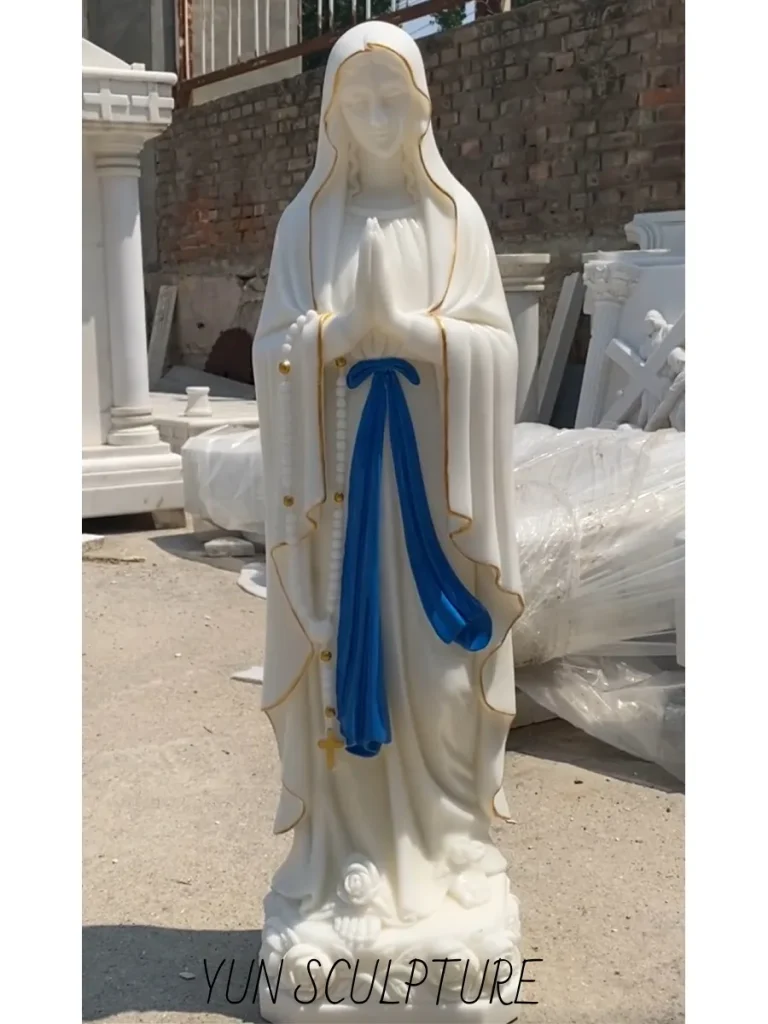 A magnificent full-length white marble Our Lady of Lourdes statue standing in a sunlit sculpture yard. Featuring the iconic blue sash and gold trim, this breathtaking artwork brilliantly demonstrates the "White Robe Theology" by allowing natural sunlight to penetrate the fine-grained stone, creating an ethereal internal glow and realistic drape that honors the 1864 Joseph Fabisch original.