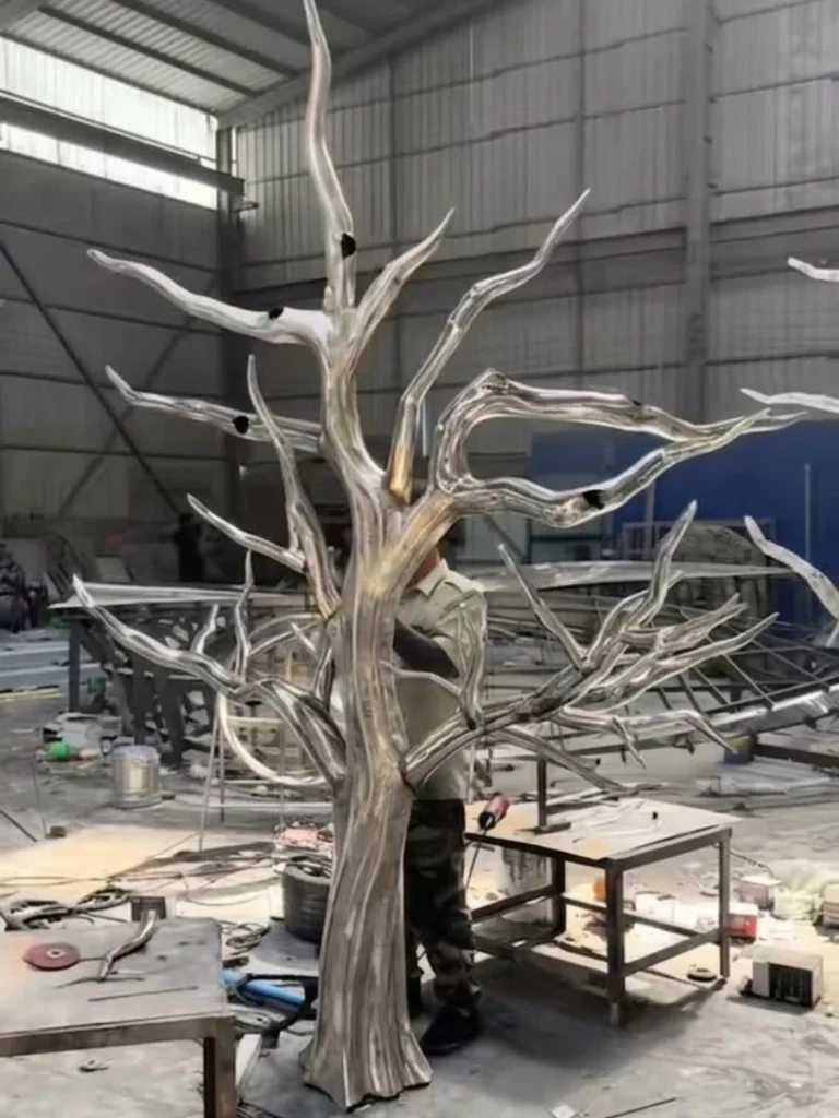 The fully clad stainless steel tree trunk and bare branches showing raw weld seams in the foundry. Hand-forged from flat metal sheets into continuous volumes, the structure achieves exceptional structural reliability and organic curvature before the intricate leaves are added.