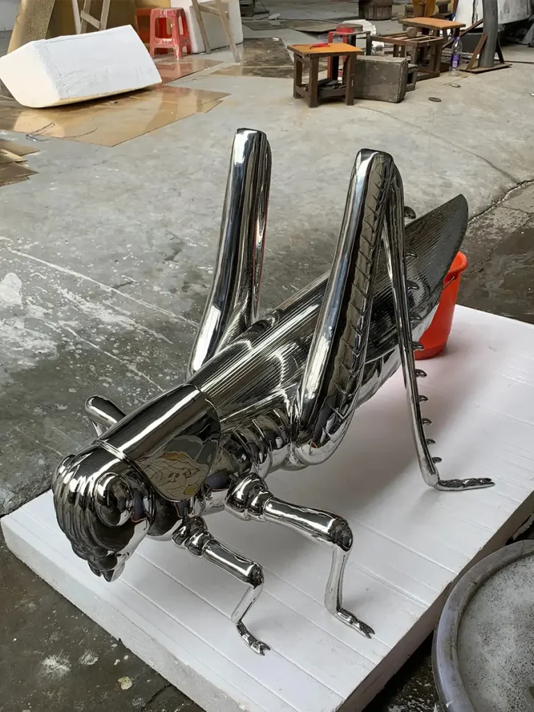 An angled view of a monumental metal grasshopper beautifully crafted from polished stainless steel. Expanding on the concept of insect art, this oversized piece translates the delicate forms of nature into a powerful architectural statement for high-end landscape design.