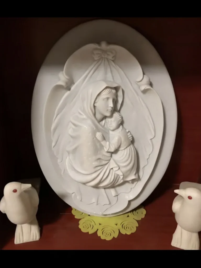 A finely carved white marble bas-relief sculpture of the Madonna and Child, displayed indoors next to two white dove figurines.