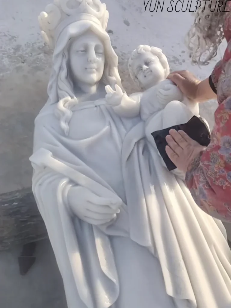 A close-up of a white marble Madonna and Child statue being hand-finished to achieve a soft, skin-like texture and translucent surface quality.