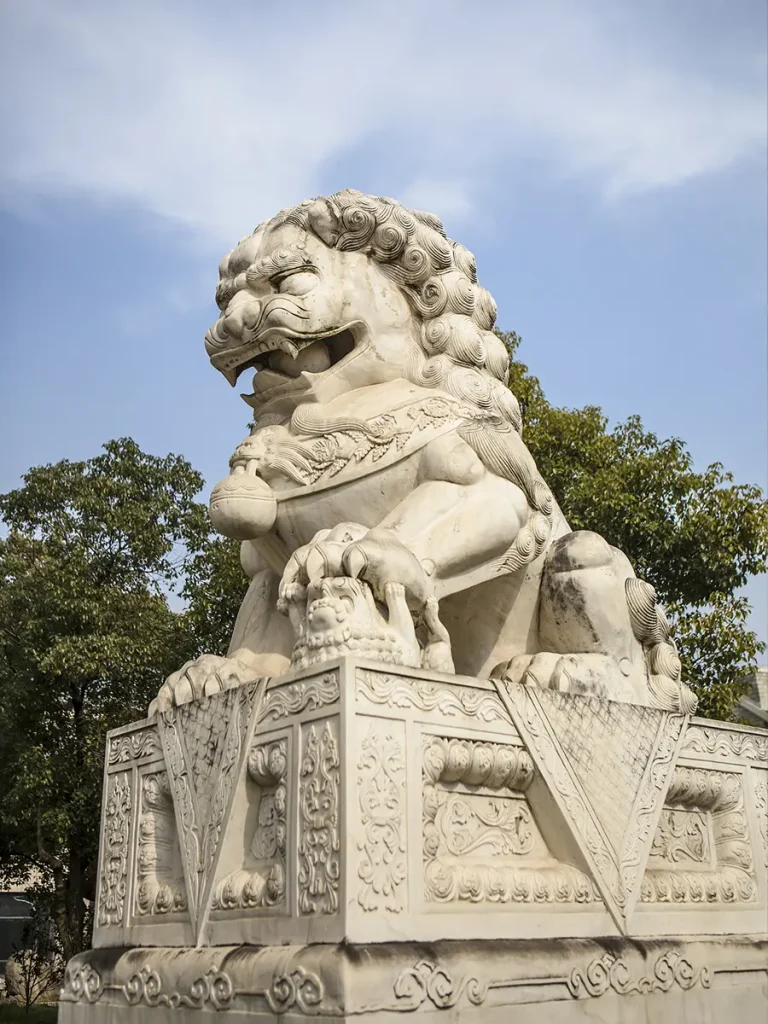 A traditional male Chinese guardian lion (Foo Dog) carved from white marble. The majestic statue rests its right front paw firmly on a decorated embroidered ball, sitting on an elaborately carved square stone pedestal with traditional drapery relief patterns, set against a blue sky.
