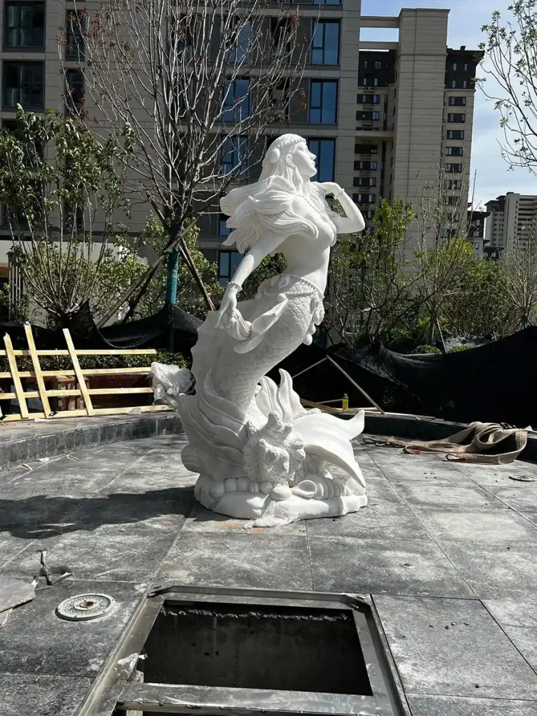 A side view of a large white marble mermaid sculpture being installed in a modern residential fountain pool, adding a mythological narrative to the landscape.