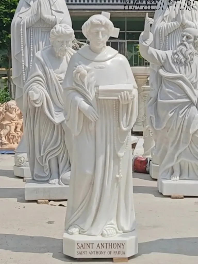 A hand-carved white marble statue of St. Anthony of Padua, featuring the saint holding a book and lily with a clearly inscribed base, crafted by Yun Sculpture.