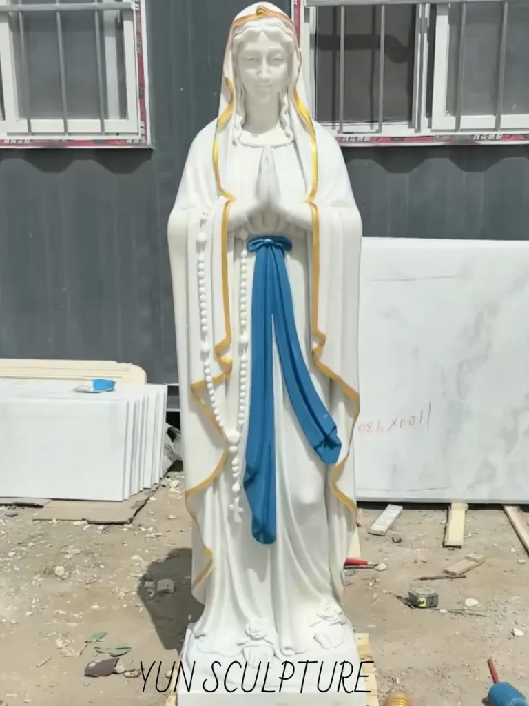 A breathtaking close-up of a white marble statue of our lady of lourdes standing in a sunlit workshop yard. Expertly hand-painted with a distinct blue sash and gold trim, this beautiful stone carving faithfully follows the permanent global reference point established by Joseph Fabisch's 1864 monumental piece, perfectly illustrating the complex technical challenge of balancing classical beauty with transcendent earthly simplicity.