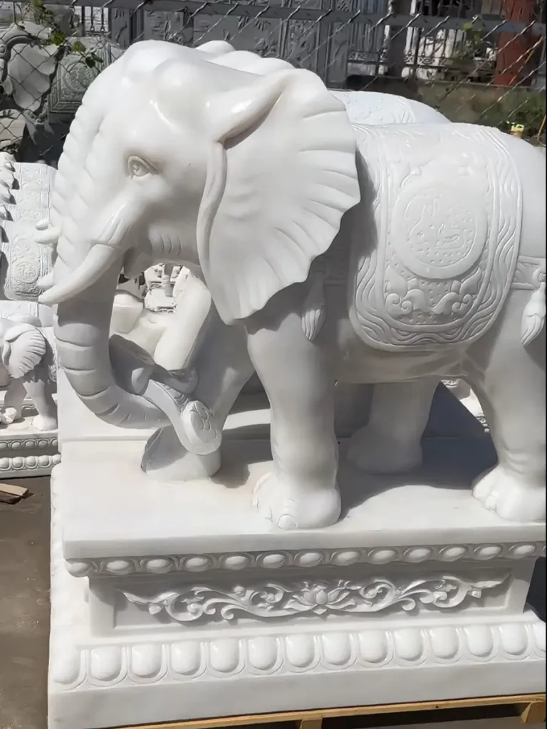 A highly detailed white marble elephant statue resting securely on a wooden shipping pallet. The extremely fine carving on its curled trunk and ceremonial blanket highlights the exceptional craftsmanship required to create these symbolic cultural guardians.