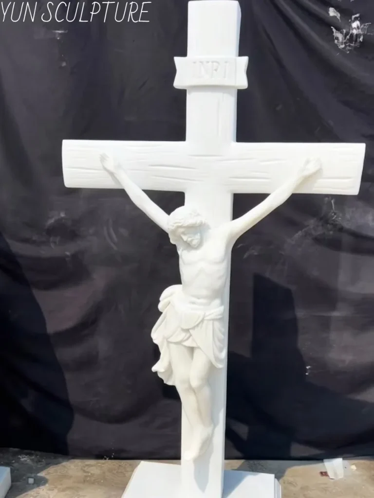 A full-length, hand-carved white marble crucifix standing against a dark backdrop. Because natural stone is vulnerable to freeze-thaw cycles and water infiltration, this majestic, dense white stone artwork is strictly specified as an altar piece for interior installation only, ensuring its pristine beauty lasts for generations.