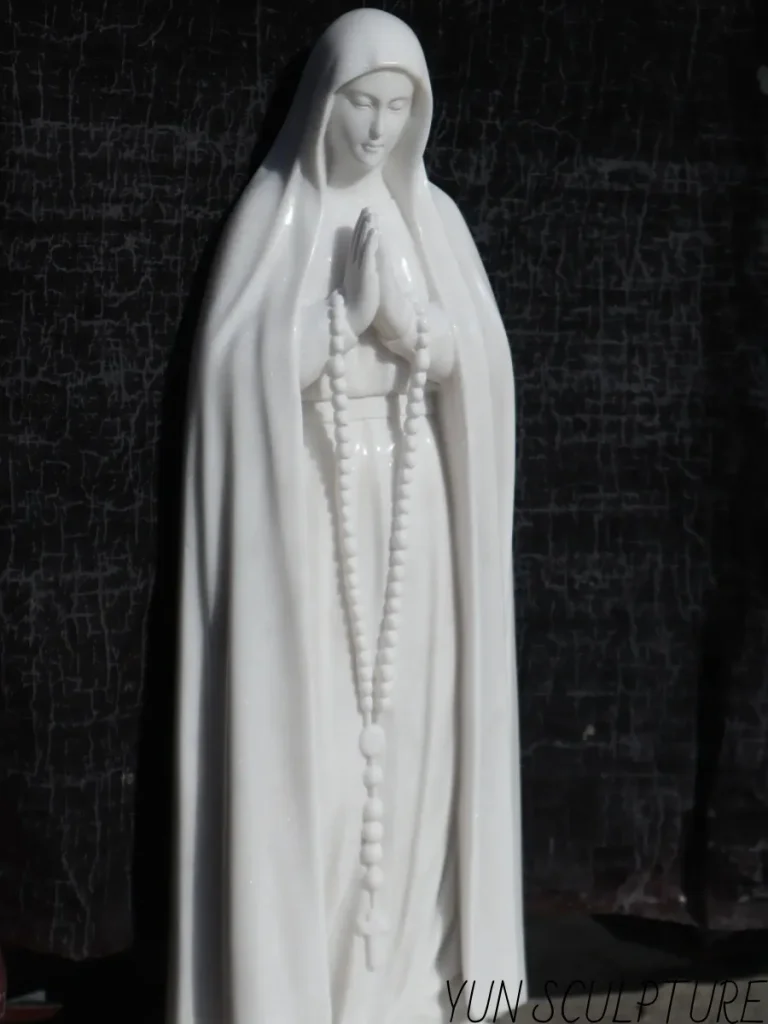 A side-angle view of a beautifully carved 5-foot white marble Virgin Mary statue standing brilliantly against a dark backdrop. Approaching life-size scale, this magnificent stone artwork ensures that parishioners can clearly see her serene facial expression and the intricate rosary details from a moderate distance, serving as a powerful public devotional presence.