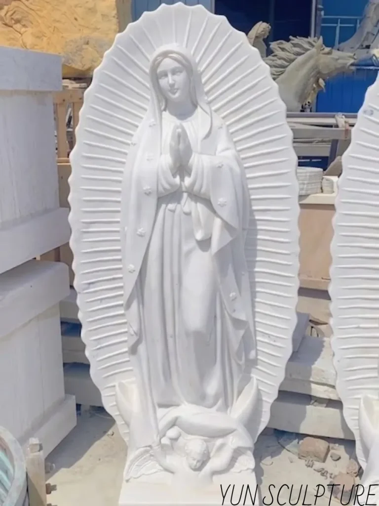 A breathtaking, full-length white marble Our Lady of Guadalupe statue standing in a sunlit sculpture workshop. Brilliantly showcasing the intense labor where master craftsmen hand-carve the sculpture from solid natural stone blocks, this monumental piece perfectly transforms the approved model into a permanent, sacred work of art capable of lasting for generations.