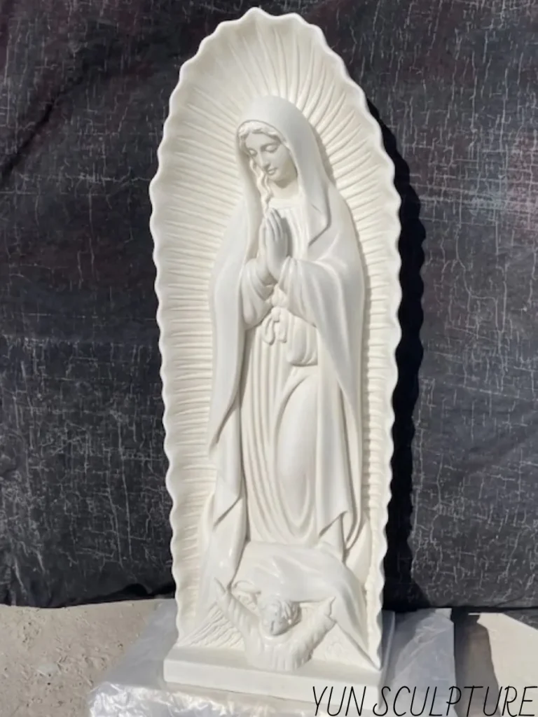 An angled full view of a pristine white stone Our Lady of Guadalupe statue resting against a dark workshop backdrop. The intricate carving of the angel and the crescent moon at her feet reveals the true devotion, masterful craftsmanship, and precise engineering required to sculpt complex religious iconography directly from solid natural marble.