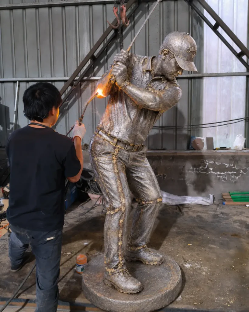 Skilled artisans welding and chasing the raw bronze segments of the golf statue, removing casting lines to create a seamless, solid metal structure.
