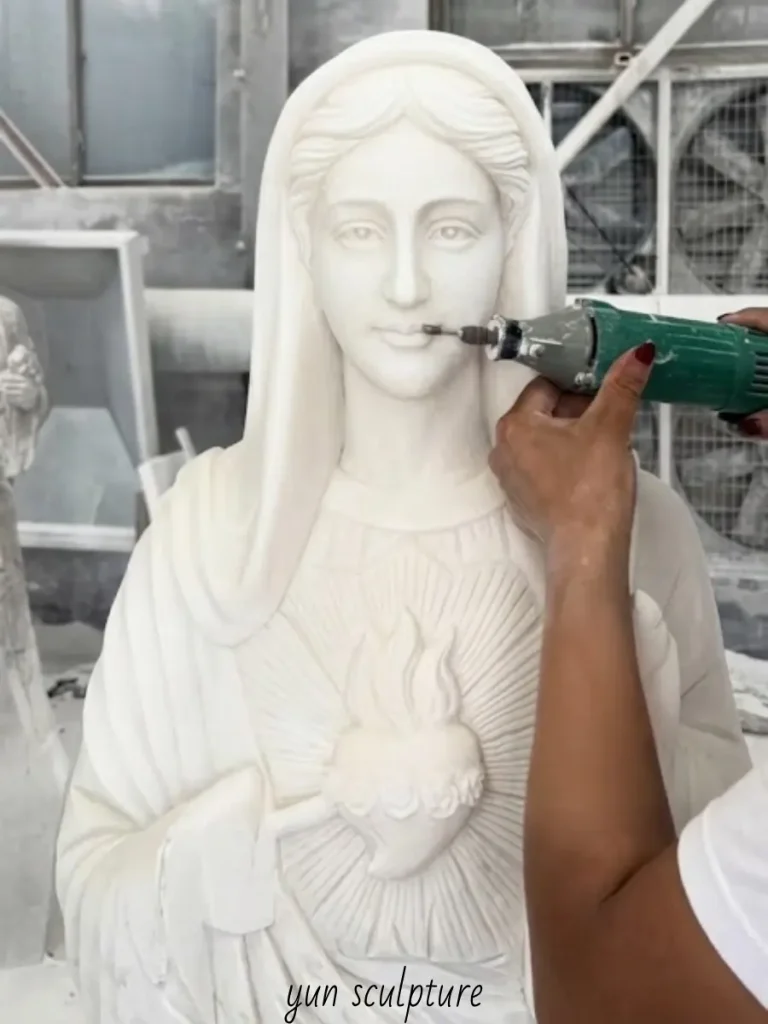 Extreme close-up of a master sculptor using a precision rotary tool to refine the delicate facial expression and the exposed heart of a marble Immaculate Heart of Mary statue. This expert craftsmanship is essential to capture the wounded peace—communicating sorrow and serenity simultaneously without collapsing into grief.