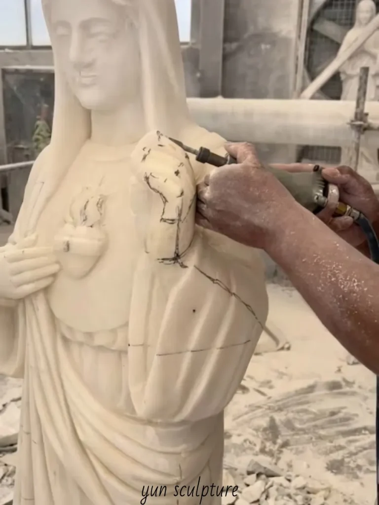 A skilled craftsman meticulously carving the flaming, pierced heart on a white marble Marian statue. This highly precise detailing visually reflects the biblical anchor of Luke 2:35—"a sword will pierce your own soul too"—manifesting her silent, enduring suffering in stone.