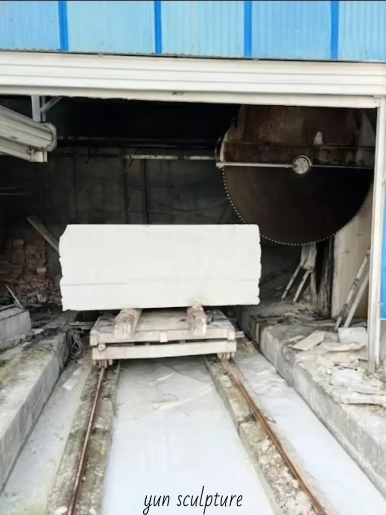 A massive raw white marble block being cut on an industrial saw, preparing to be transformed into a profound theological statement—a custom Immaculate Heart of Mary statue representing love that suffers without retreating.