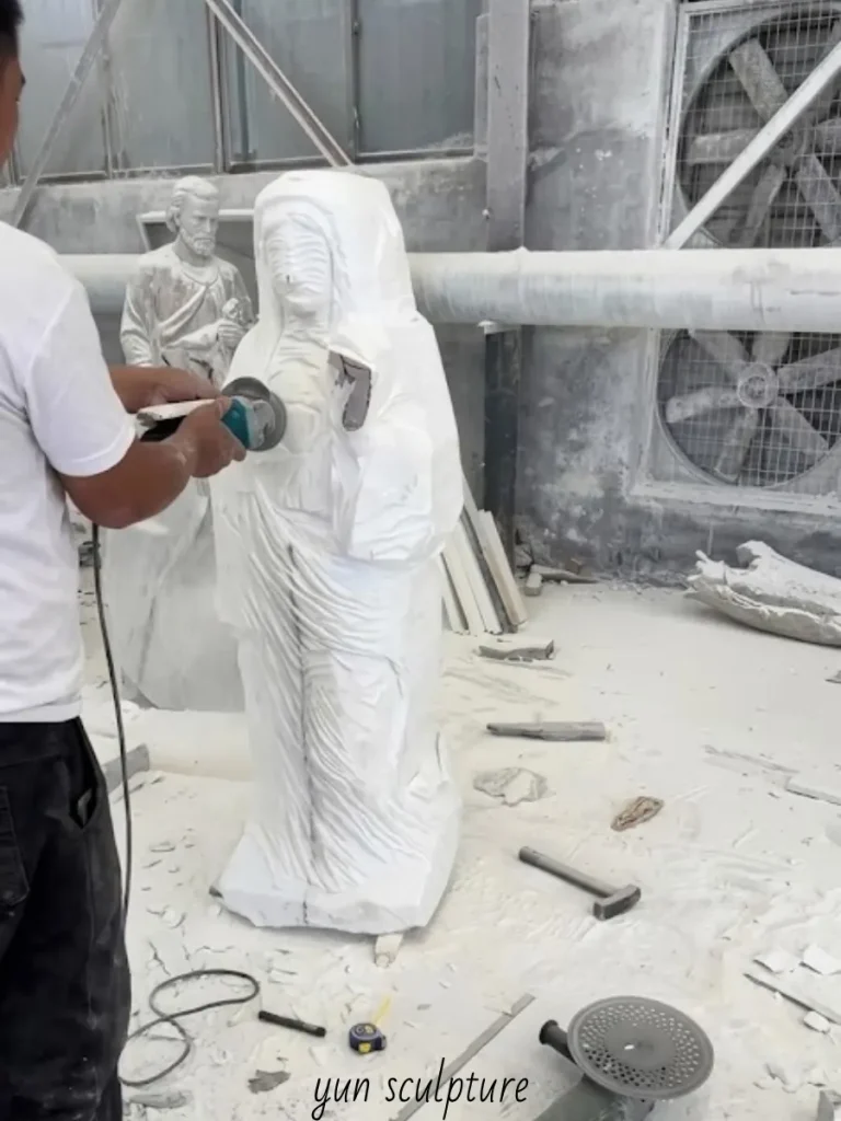 An artisan using a grinder to shape the flowing robes and receptive posture of a white marble Virgin Mary statue, slowly bringing her complete, unguarded self-giving out of the solid rock.