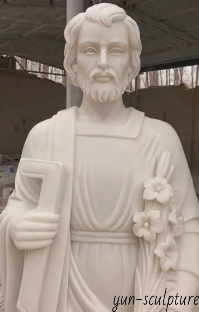 Close-up detail of a high-quality marble St. Joseph statue, showing the intricate carving of his face, carpenter's square, and lilies, designed as a permanent garden fixture.