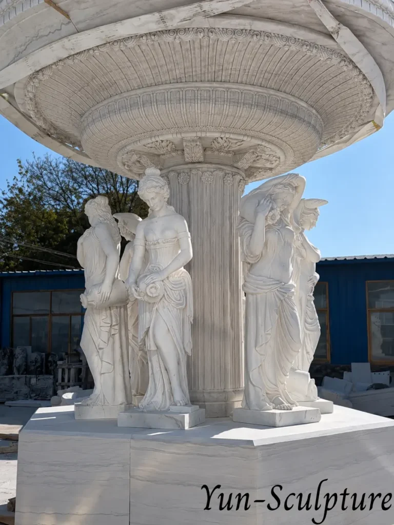 The base of a grand white marble fountain featuring a group of Venus-inspired figures. This design follows the practical rule of garden placement, where each figure has a clear direction of arrival, facing the primary viewing point while the water feature remains behind them.