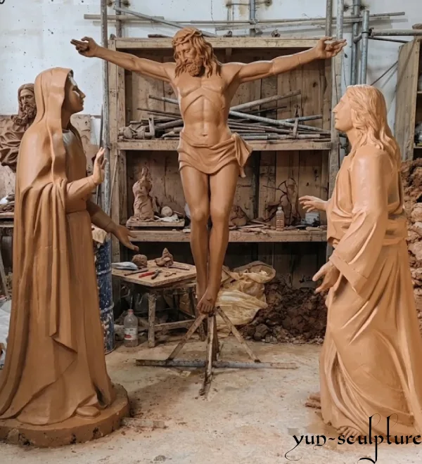 A 1:1 scale clay model of a Crucifixion group sculpture, featuring Jesus on the cross flanked by Mary and Saint John, being hand-sculpted in our workshop. This stage ensures the spiritual meaning and anatomical accuracy of our custom liturgical art pieces before casting.