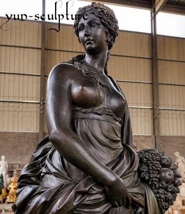 A promotional card on a blue background featuring a photo of a classical dark bronze sculpture of a female deity representing Venus or Aphrodite. The figure has styled curly hair, wears draped classical robes, and stands next to a cornucopia filled with grapes and other fruits. A stylized black "yun-sculpture" watermark is visible in the top left corner. Below the photo, the text reads: "Venus & Aphrodite - The Goddesses of Beauty and Nature. Perfect for gardens and courtyards."